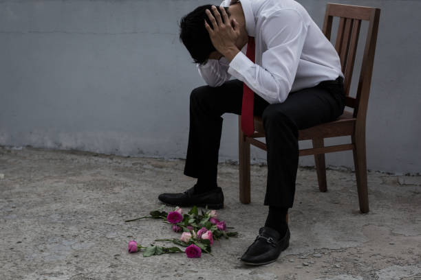 Sad brokenhearted man rejected after a date. A man dressed in a suit sitting on a chair with roses lying on the ground, sad from his heart being broken, dramatic setting. face down stock pictures, royalty-free photos & images