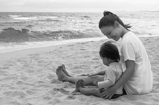 A mother holding her young toddler sitting together on the sand near the water. Happy family.
