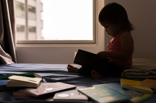 Little genius girl fascinated with books, sitting on her bed reading a story near the window. Bookworm.