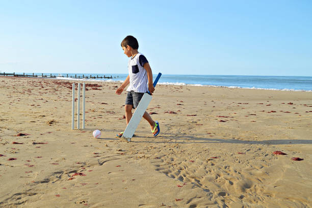 petit garçon restant sur une plage retenant une chauve-souris de cricket, regardant les guichets - sport of cricket cricket player cricket bat batting photos et images de collection