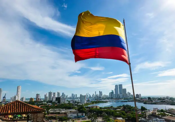 The Colombian flag waving in the wind. Blue sky with white clouds in the background. The city of Cartagena is in the far back. Shot in Cartagena, Colombia, USA.