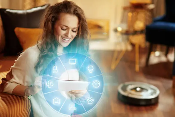 Photo of housewife using smart home application on tablet PC and robot vacuum cleaning floor in background