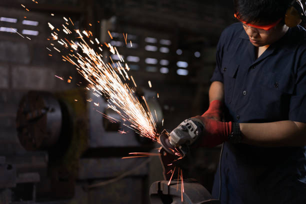 ethnic asian workshop metal worker wearing safety equipment and operating an angle grinding machine on steel causing metal sparks fly - grinding grinder work tool power tool imagens e fotografias de stock