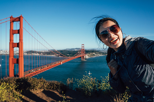 Happy young female tourists wearing sunglasses taking selfie in San Francisco by Golden Gate Bridge USA. asian woman face camera smiling attractive taking self portrait with famous attraction in us.