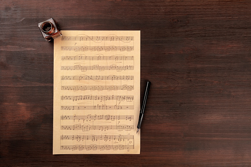 An overhead photo of a piece of sheet music, shot from the top on a dark rustic wooden background with an ink well and a nib pen, with a place for text