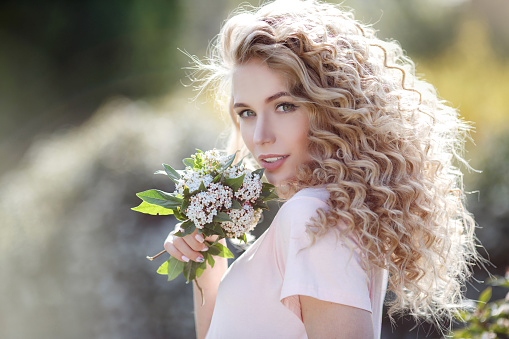 Beautiful young model spring girl in flowers with hairstyle in summer blossom park. Woman in a blooming garden . Fashion, Cosmetics & Perfumes.Portrait of a beautiful woman with curly hair outdoors in a spring park