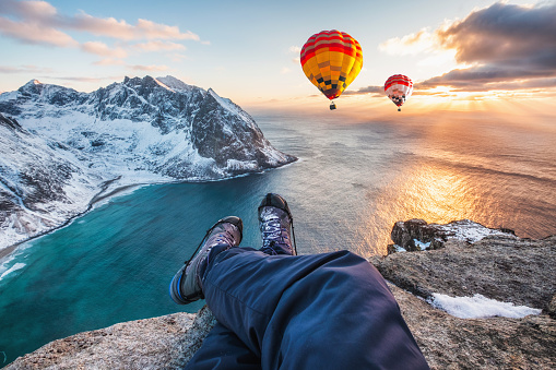 Man hiker cross legs sitting on rock ridge with hot air balloon flying on ocean at sunset