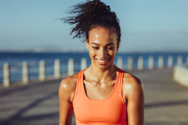 Sporty woman taking a break while exercising Smiling young female runner taking a breather. Healthy young woman with sweat standing on the promenade after her workout and smiling. healthy lifestyle women outdoors athlete stock pictures, royalty-free photos & images