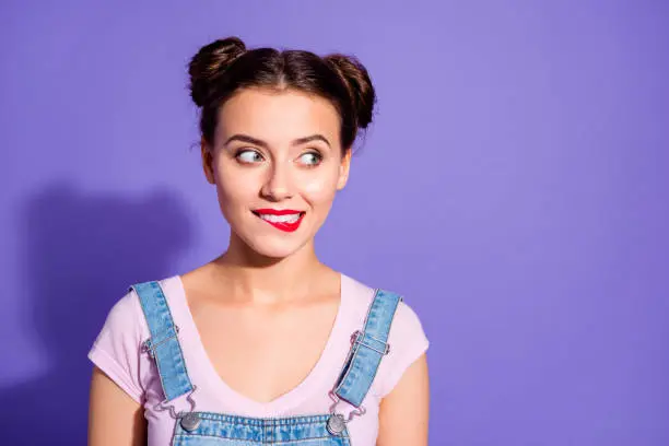 Close up photo beautiful amazing she her lady two buns red pomade bite upper lip tricky mood look side empty space wear casual t-shirt jeans denim overalls clothes isolated purple violet background