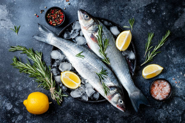 pescado fresco de la lubina cruda sobre fondo de piedra negra con especias, hierbas, limón. fondo culinario de marisco con ingredientes para cocinar. vista superior - fish fotografías e imágenes de stock