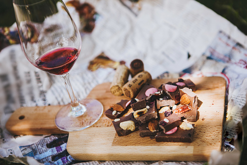 Glass with red wine and pieces of chocolate with nuts and raisins stands on cutting board on background of newspapers on the ground outdoors. Alcoholic drink in glassware with snacks