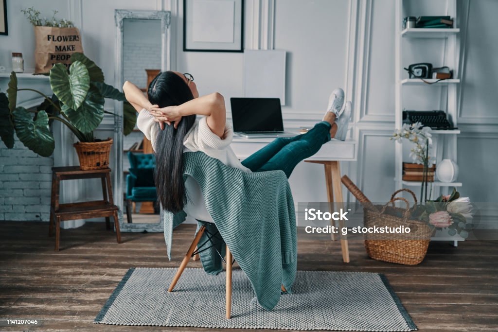 Total relaxation. Rear view of young woman keeping hands behind head while sitting in home office Feet Up Stock Photo