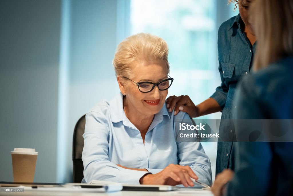 Expertise discussing new plans with businesswomen Smiling elderly expertise discussing plans with businesswomen. Professionals are building new strategy together. They are working in start-up office. 35-39 Years Stock Photo