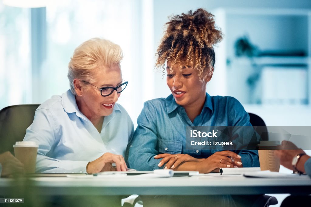 Smiling businesswomen working in new office Smiling businesswomen working at desk in new office. Confident female executives are planning strategies. Senior expertise is advising colleague. Advice Stock Photo