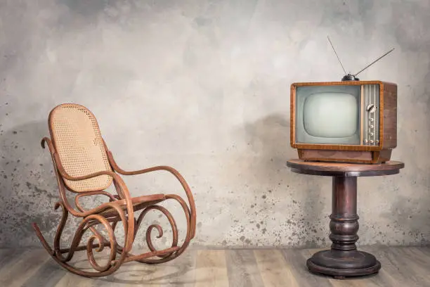 Photo of Retro old outdated TV receiver from circa 50s on wooden table and aged rocking chair front textured concrete wall background. Vintage style filtered photo