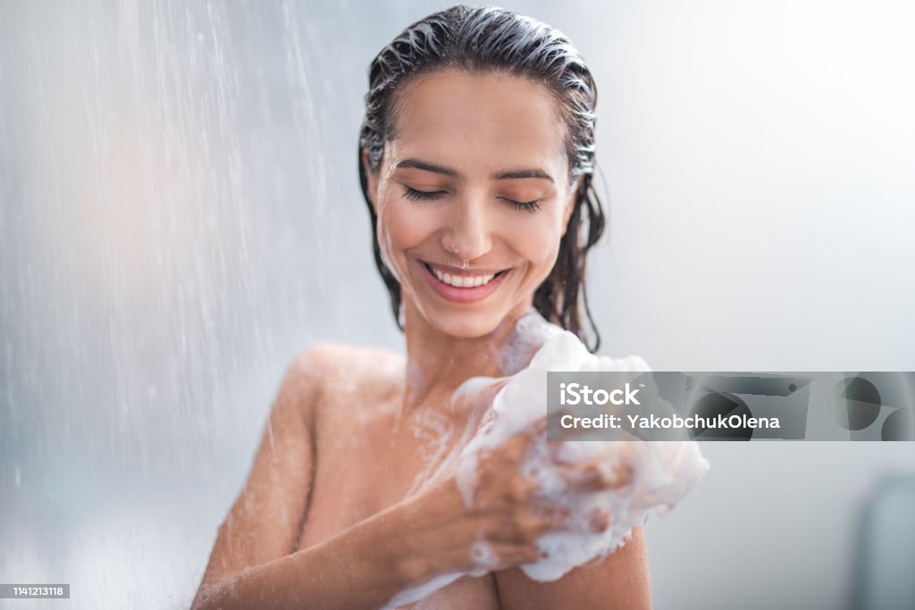 Smiling female rubbing body with foam Portrait of happy girl taking shower with gel. She washing with puff Shower Stock Photo