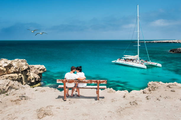 Honeymoon traveller couple hugging on a wooden bench and enjoys their tropical holiday. Wedding travel. Young happy couple sitting back on beach. Rear view of a couple in love on vacation. Travelers stock photo
