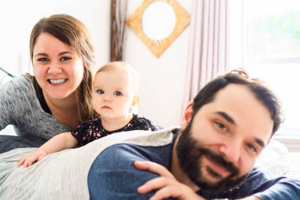 une petite fille sur le dos de son père dans la literie blanche à la maison avec la mère - baby beauty beautiful the human body photos et images de collection