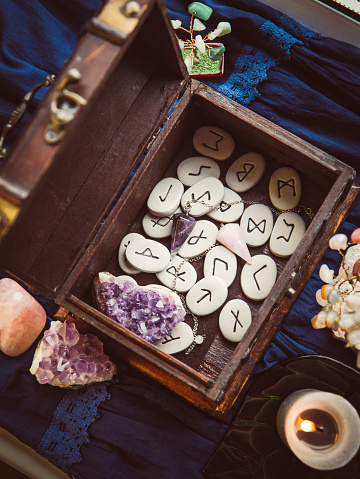 Clairvoyant tools rune stones, crystal pendulums in natural dark wooden case box on dark blue background. Instagram filter.