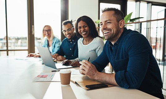 Diverse group of young businesspeople laughing while sitting together in a row at an office desk during a meeting