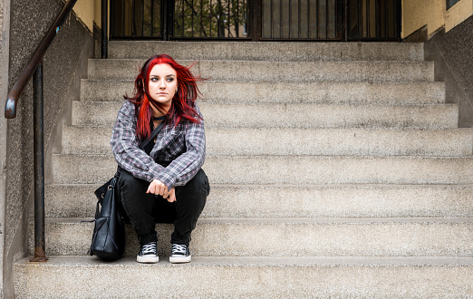 Homeless girl, Young beautiful red hair girl sitting alone outdoors on the stairs of the building with hat and shirt feeling anxious and depressed after she became a homeless person
