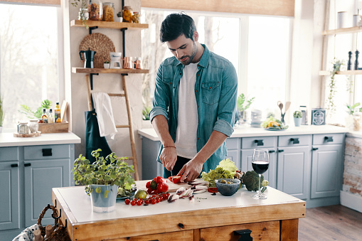 Handsome young man in casual wear cutting vegetables while standing in the kitchen at home