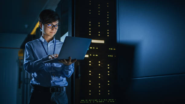 w dark data center: mężczyzna specjalista it stoi obok rzędu operacyjnych stojaków serwerowych, używa laptopa do konserwacji. koncepcja chmury obliczeniowej, sztucznej inteligencji, superkomputera, cyberbezpieczeństwa. neonowe - network server rack data center in a row zdjęcia i obrazy z banku zdjęć