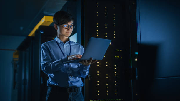 w dark data center: mężczyzna specjalista it stoi obok rzędu operacyjnych stojaków serwerowych, używa laptopa do konserwacji. koncepcja chmury obliczeniowej, sztucznej inteligencji, superkomputera, cyberbezpieczeństwa. neonowe - network server rack data center in a row zdjęcia i obrazy z banku zdjęć