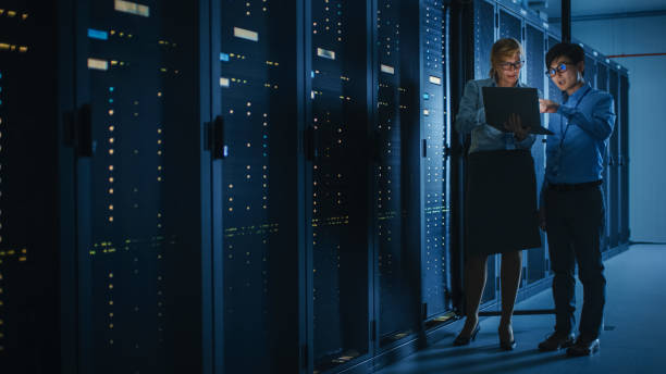 In the Modern Data Center: Engineer and IT Specialist Work with Server Racks, Talking, Doing System Maintenance Check with Laptop Computer. Concept of Diagnostics of the Database, Cybersecurity Check en el centro de datos moderno: ingeniero y especialista en ti trabajar con bastidores de servidor, hablar, hacer comprobación de mantenimiento del sistema con el ordenador portátil. concepto de diagnóstico de la base de datos, verificación de cibersegu