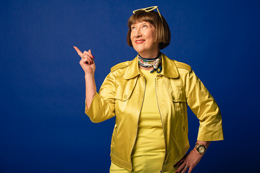 Portrait of a smiling mature woman dressed in a quirky gold outfit looking up while she points in front of a dark blue background.
