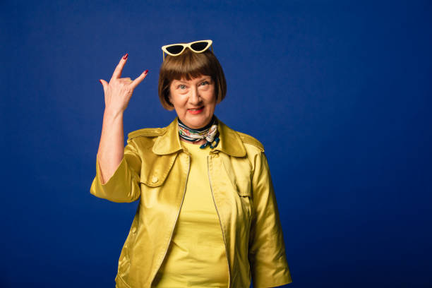 Rock And Roll Portrait of a smiling mature woman dressed in a quirky gold outfit in front of a dark blue background. She is doing the sign of the horns, also known as the 'rock and roll' sign. horn sign stock pictures, royalty-free photos & images