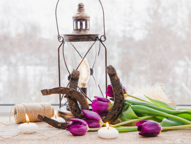 istock Old rusty horseshoes, brown vintage lantern with modern dimmable decorative light bulb switched on and white candles lit and  tulips on wooden window sill. Horseshoe Vastu Shastra good luck symbol. 1141230582