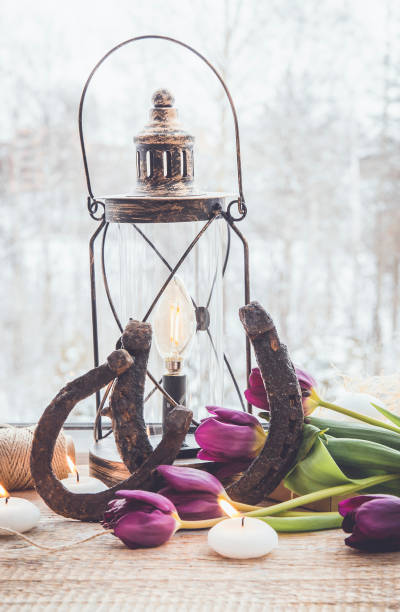 Old rusty horseshoes, brown vintage lantern with modern dimmable decorative light bulb switched on and white candles lit and  tulips on wooden window sill. Horseshoe Vastu Shastra good luck symbol.