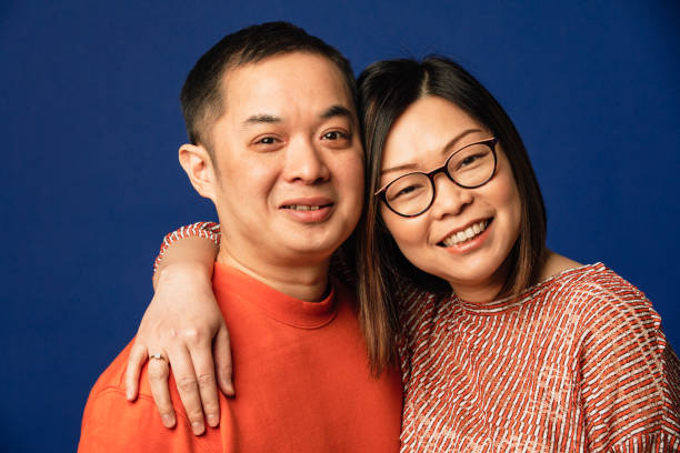Close up portrait of a couple of Chinese ethnicity side by side with their arms around each other looking at the camera as they smile. They are standing in front of a blue coloured background.