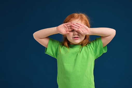 Funny photo of little redhead girl covering her eyes up from bright light or sun, blue background, green t-shirt