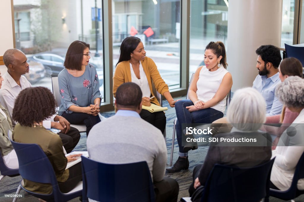 Mental health professional comforts woman Upset mid adult woman discusses a difficult issue during a support group meeting. A caring mental health professional comforts and speaks encouraging words to the woman. Group Therapy Stock Photo