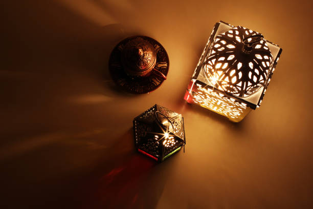 Festive table composition of glowing Moroccan ornamental lanterns and bronze tea cup. Decorative golden shadows. Greeting card, invitation for Muslim holy month Ramadan Kareem, flat lay, top view.
