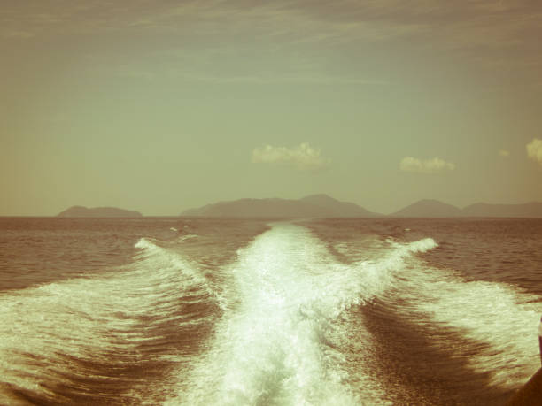 A boat sailing on transparent blue sea against mountains and blue sky away from the Surin Islands in the Andaman Sea. They are famous for their reefs and snorkeling. This is  a view from the rear of the speedboat with the wake from its 3 engines in the foreground.