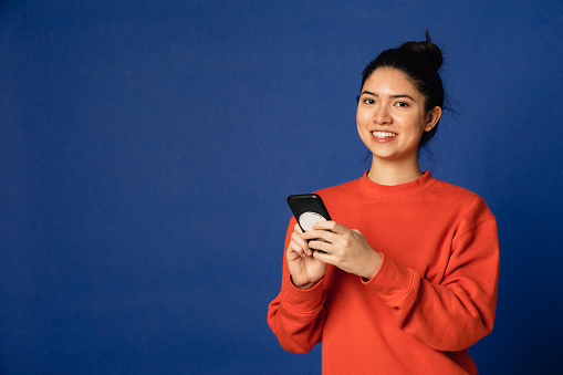 Portrait of a woman in her twenties dressed casually in front of a blue studio background holding a mobile phone.