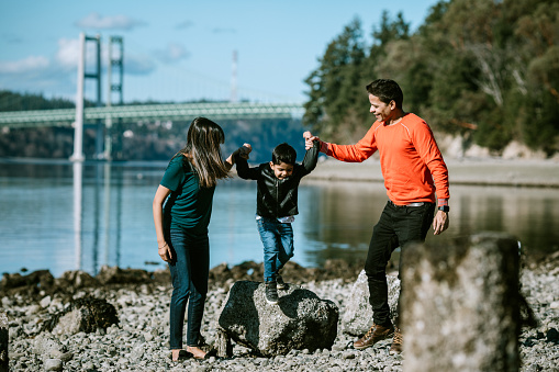 A young family spends time together at a Puget Sound beach in Washington state, enjoying the time outdoors with each other.  The father and mother carry their little boy in their arms.