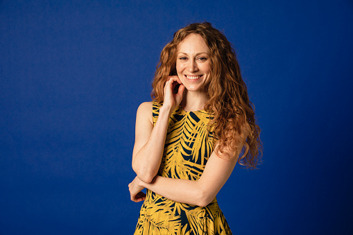 Portrait of a woman smiling as she looks at the camera in front of a blue studio background. Her arms are crossed and she has one hand on her chin.