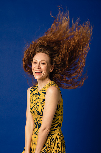 Portrait of a woman laughing as she looks at the camera tossing her hair. She is standing in front of a blue studio background.