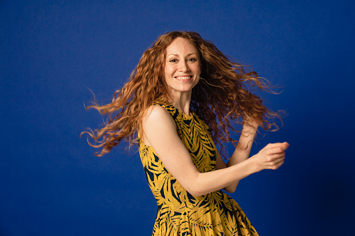 Portrait of a woman smiling as she looks at the camera tossing her hair. She is standing in front of a blue studio background.