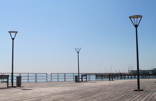 Empty wooden sea pier on Limassol Molos promenade with metal railings, street lights, benches and litter bins in front of the blue sky on a sunny spring day