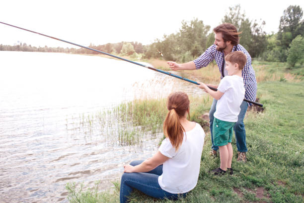 istock Guy is teaching his son how to catch fish right. Boy is holding long fish-rod. Guy is guide him. Young woman is sitting close to them. 1141251622