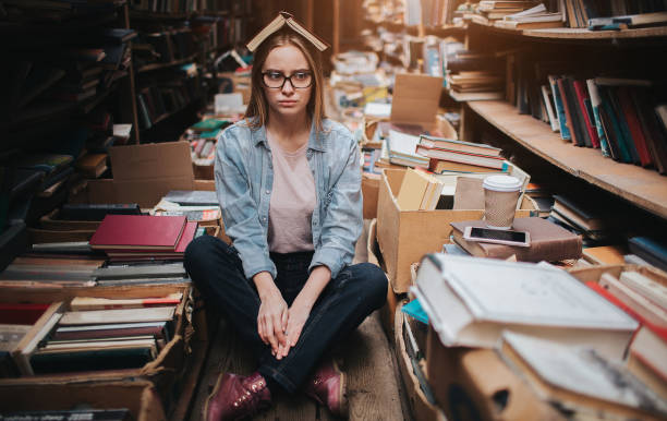 Tired and sad teenager is sitting on the floor among many books. There is also an opened book on her head. This girl didn't find any information for her homework. She is upset stock photo