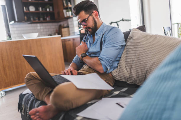 istock Modern bearded young businessman working on laptop computer while sitting on sofa at home, looking at some documents 1141258999