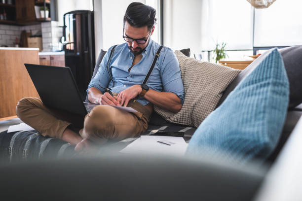 istock Modern bearded young businessman working on laptop computer while sitting on sofa at home, looking at some documents 1141259075