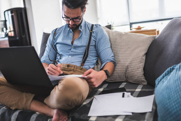 istock Modern bearded young businessman working on laptop computer while sitting on sofa at home, looking at some documents 1141259399