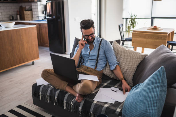 istock Modern bearded young businessman working on laptop computer while sitting on sofa at home, looking at some documents 1141259402